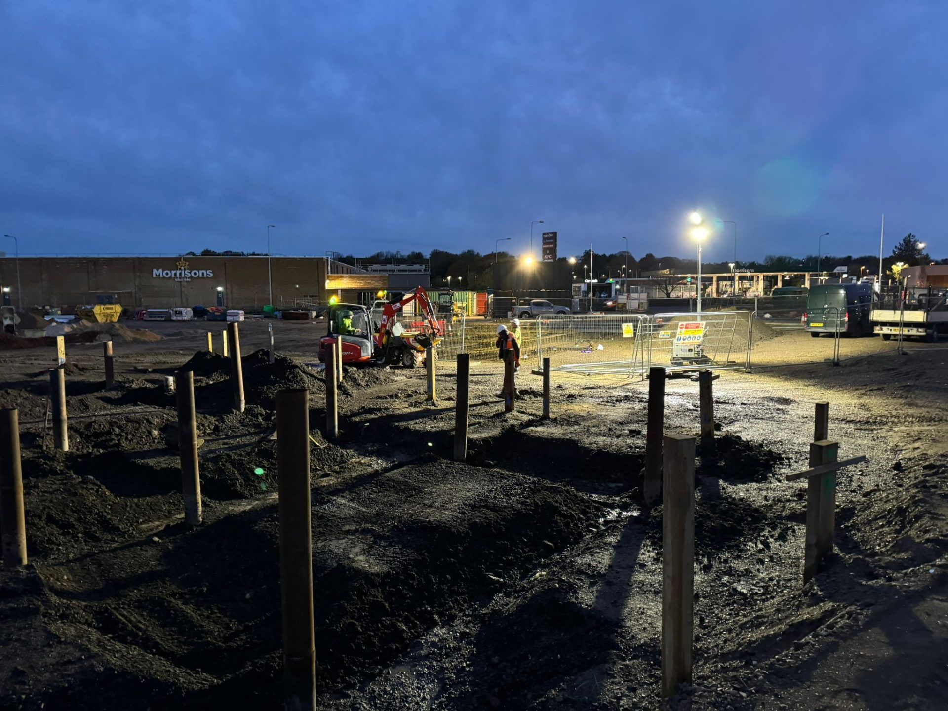 Floodlit view of groundworks with mini digger and several workers.