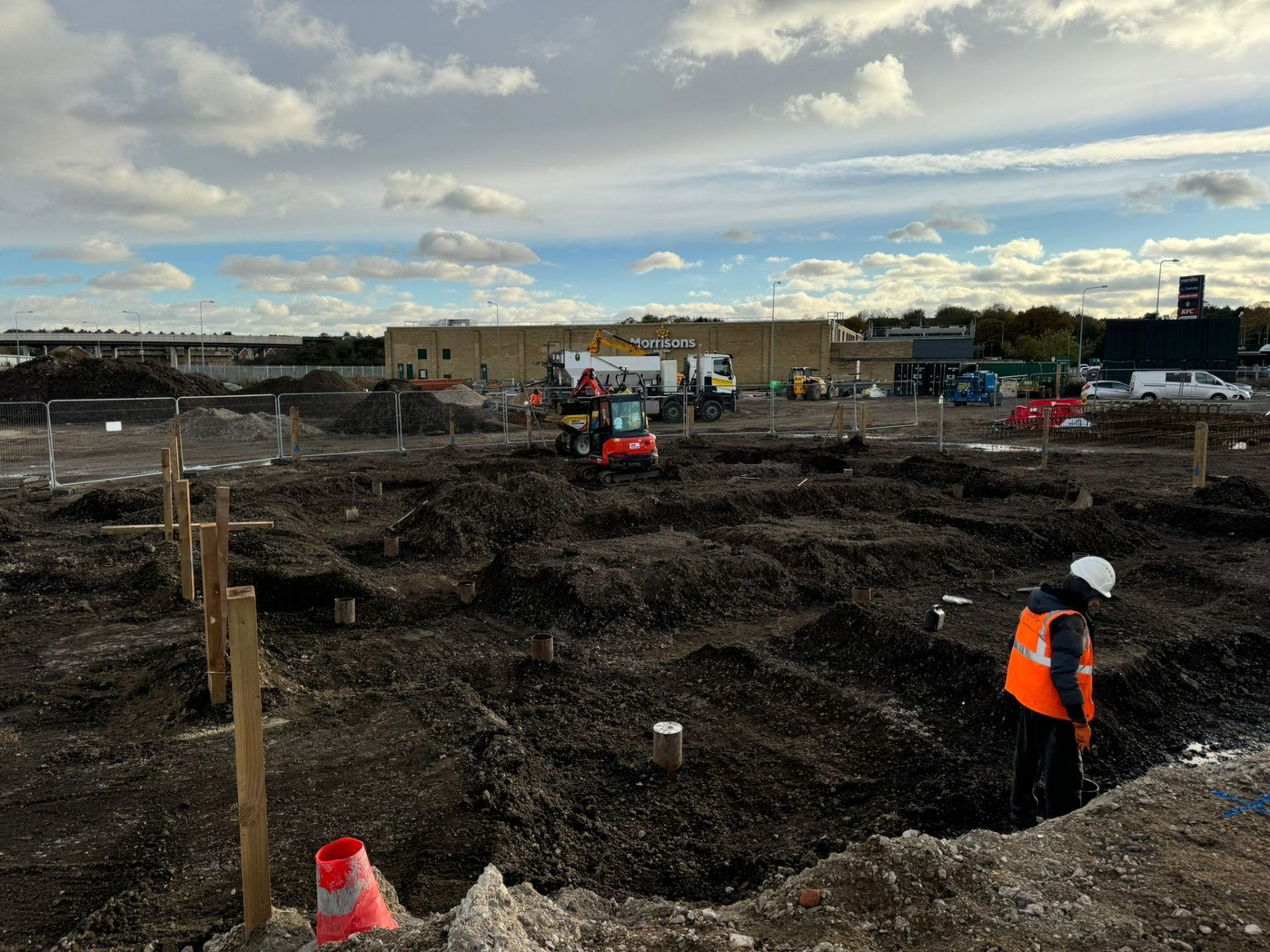 Excavated groundworks with workman in hard hat in foreground and red mini digger in background.